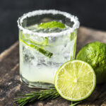 Image shows a highball glass rimmed with salt, filled with white soda, mint and limes. A sliced lime sits next to the glass on a table with more garnish.