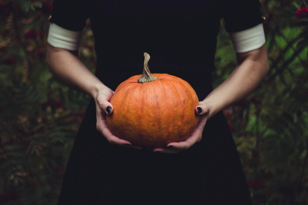 Image shows the torso of a woman dressed in black holding an orange pumpkin.