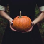 Image shows the torso of a woman dressed in black holding an orange pumpkin.